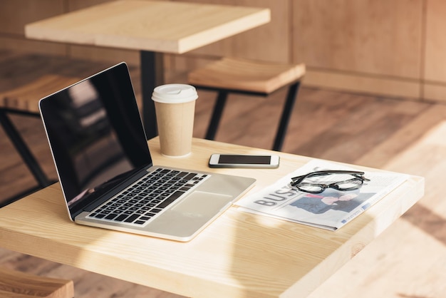 Laptop mit leerem Bildschirm, Smartphone und Wirtschaftszeitung auf dem Tisch im Café