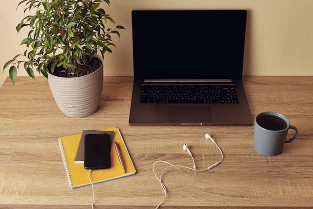 Laptop mit leerem Bildschirm, Topfpflanze, Becher, gelbem Notizblock, Stift, Handy und Kopfhörern.