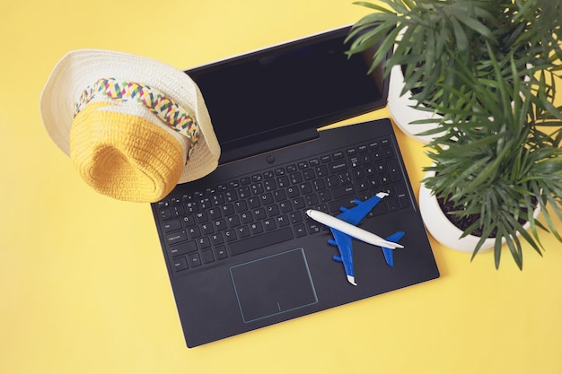 Laptop, Modellflugzeug, Strohhut und Palmblätter auf gelbem Hintergrund. Sommerurlaub, Urlaub, Online-Buchung, Reisekonzept planen. Flache Lage, Ansicht von oben.