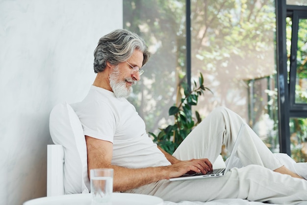 Laptop nas pernas homem moderno elegante sênior com cabelos grisalhos e barba dentro de casa