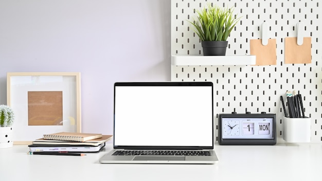 Laptop, pegboard e material de escritório na tabela de espaço de trabalho com computador monitor vazio.