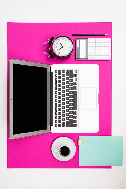 Laptop, Tasse, Uhr, Notizbuch liegen an einer rosa Wand