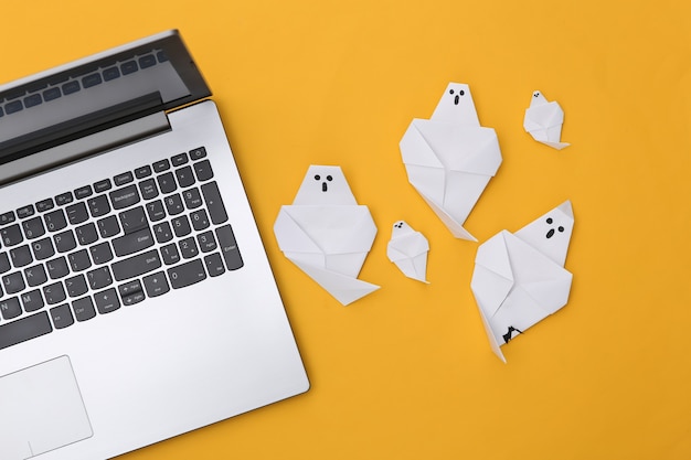 Laptop- und Origami-Geister auf gelbem Hintergrund. Halloween-Thema. Ansicht von oben