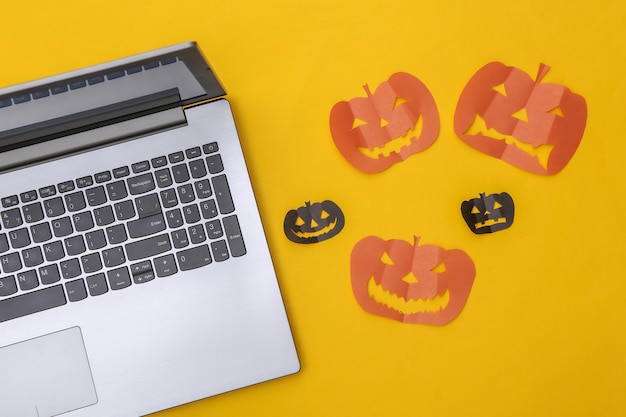 Laptop und Scherenschnitt Halloween-Kürbisse auf gelbem Hintergrund. Halloween-Thema. Ansicht von oben