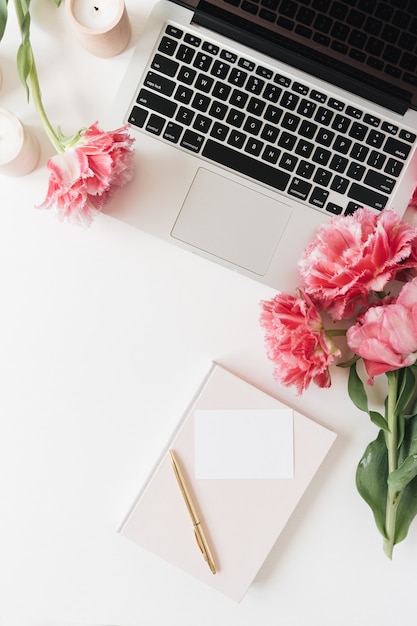 Laptop und schöne rosa Pfingstrosen-Tulpenblumen, leere Papierblattkarte, Kerzen auf Weiß