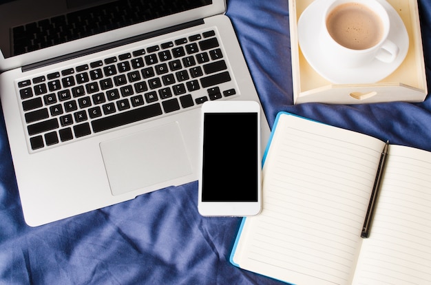 Laptop und Smartphone, Kaffeetasse und Notebook auf dem Bett in der Morgenzeit. Attrappe, Lehrmodell, Simulation.