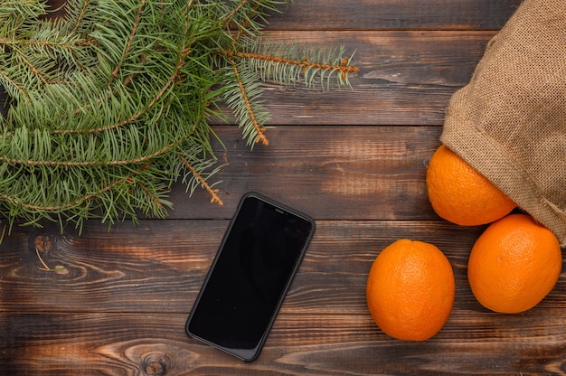 Laranjas em um saco de linho em uma superfície de madeira perto de ramos de pinheiro e smartphone preto natal