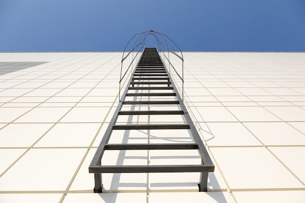 Una larga escalera de metal en la moderna fachada gris de un edificio industrial, almacén o centro comercial contra el cielo azul. Símbolo de la escalera al cielo. Concepto de equipo de extinción de incendios