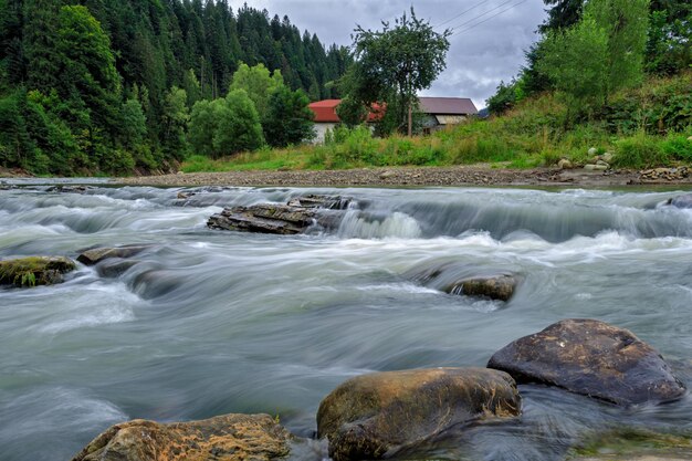 Larga exposición de un río de montaña con rocas desde el fondo de la toma con árboles y una casa en el...