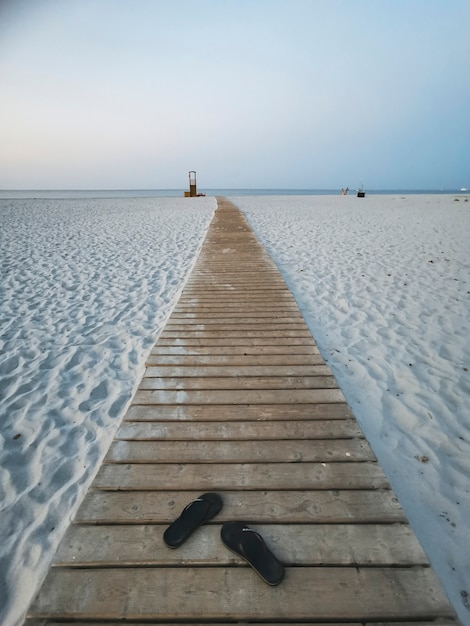 Largo camino de madera en la arena de la playa blanca al agua de mar con un par de sandalias solo - concepto de personas y vacaciones de verano - vista al mar y al cielo en backgorund