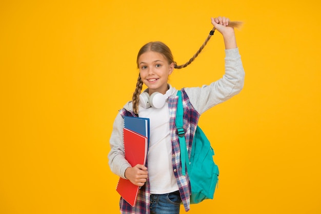 Largo y trenzado Lindo pequeño escolar sostiene una trenza de cabello largo sobre fondo amarillo Adorable niña usa un estilo de cabello largo Peinado de cabello largo para la escuela