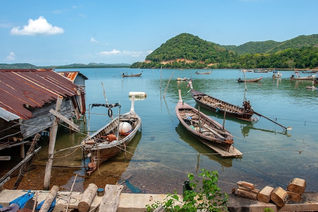 Largos barcos de pesca en Tailandia en el agua junto a la cabaña sobre pilotes en el agua con techo oxidado viejo