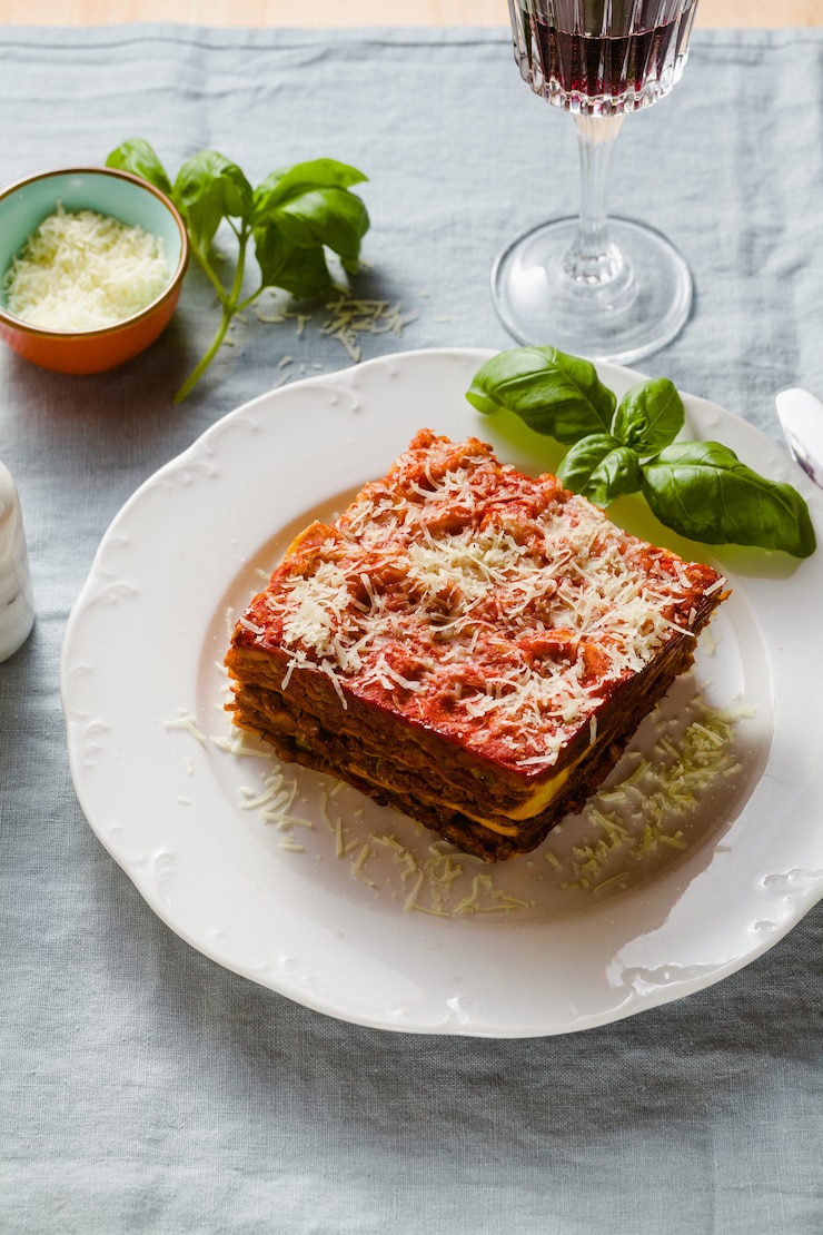 Foto lasanha vegana com lentilhas e ervilhas verdes em uma assadeira sobre uma mesa com uma toalha de mesa de linho azul. e vinho tinto em copos