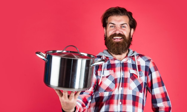 Lass uns essen. Mann hält Bratpfanne .. männlicher Küchenchef Kitchener Kochen. Hipster kariertes Hemd für bärtige Kerlkoch. unrasierter brutaler Mann mit Küchengerät. Haushaltsgegenstände. freundliche Verkäuferin.