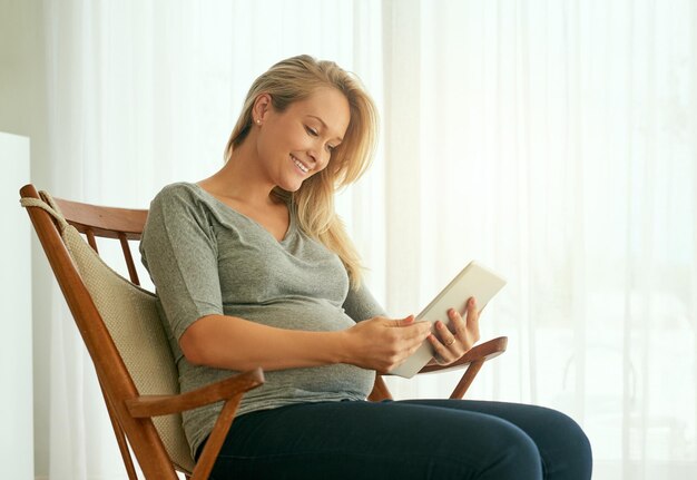 Lassen Sie sich von der Technologie durch den Prozess leiten Aufnahme einer schwangeren Frau, die ihr digitales Tablet verwendet, während sie auf einem Schaukelstuhl sitzt