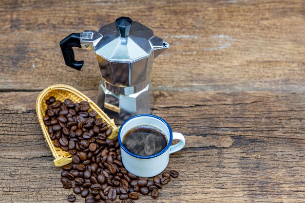 Una lata blanca taza de café caliente con cafetera italiana (Moka) y granos de café en la mesa de madera.