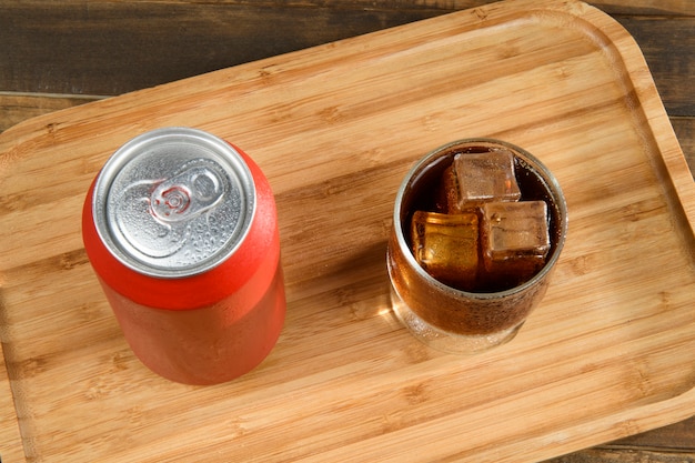 Foto lata de refresco con un vaso lleno de hielo y bebida en bandeja de bambú