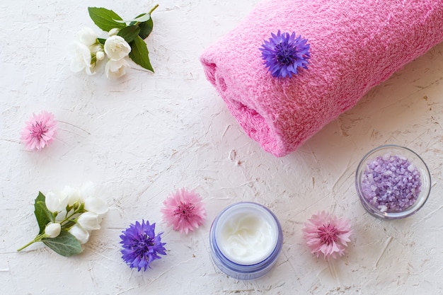 Latas com sal marinho e creme para a pele, toalha e flores de jasmim e centáurea em um fundo branco. Produtos femininos para spa. Vista do topo.