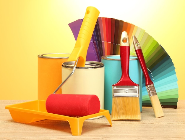 Latas de estaño con pinceles de pintura y paleta de colores brillantes sobre una mesa de madera sobre fondo amarillo