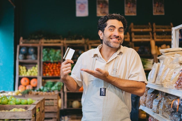 Lateinamerikaner mittleren Alters zeigt auf eine Kreditkarte, während er in einem Bioladen die Schultern hebt. Kopierraum