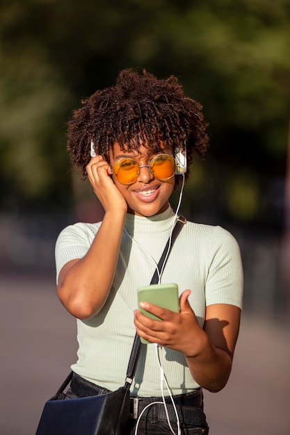 Lateinamerikanische junge Frau mit Afro-Haaren und Brille lächelt glücklich mit Smartphone und Kopfhörern in der Stadt