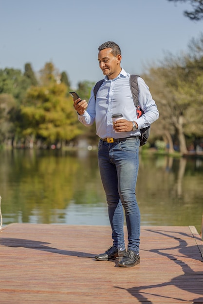 Lateinamerikanischer Mann auf einem Pier mit einer Einwegtasse Kaffee in der Hand und Blick auf sein Mobiltelefon
