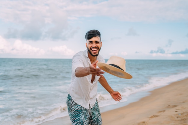 Lateinamerikanischer Mann, der lächelt und seinen Hut wirft, der die Kamera am Strand an einem schönen Sommertag betrachtet