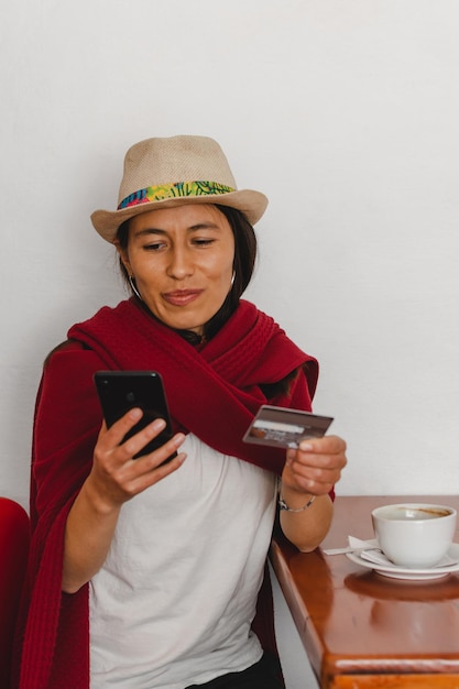 Lateinische Bäuerin mit Hut und rotem Schal, die ein Handy und eine Kreditkarte in den Händen hält Elektronisches Zahlungskonzept
