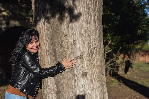 Lateinische erwachsene Frau im Freien, die die Kamera anschaut und einen großen Baum umarmt