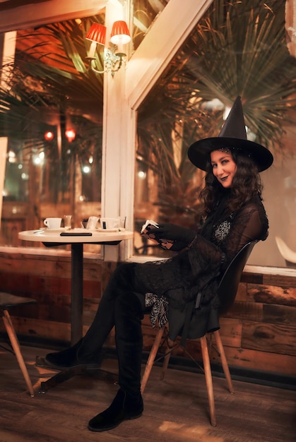 Lateinische Frau, die als Hexe verkleidet ist und in einem Café mit einem Buch in den Händen sitzt und böse lächelt