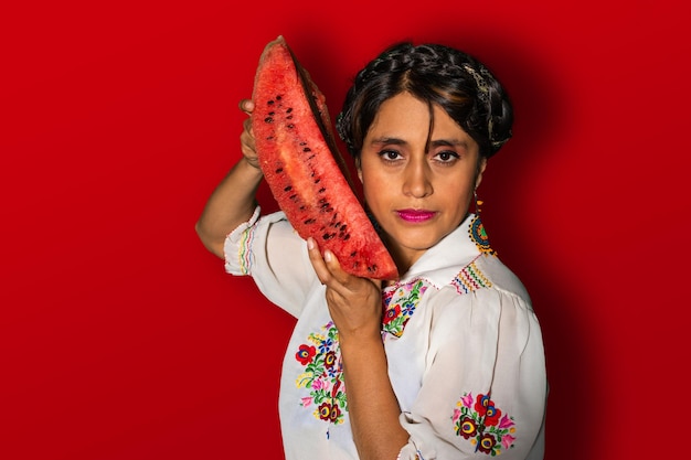 Lateinische Frau in traditioneller Kleidung, die ein Stück Wassermelone neben ihrem Gesicht auf rotem Hintergrund hält