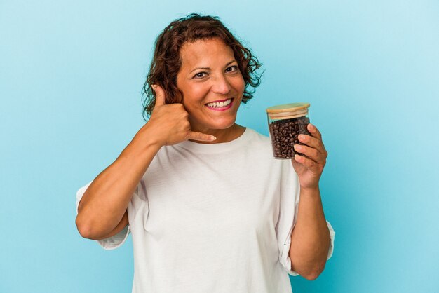 Lateinische Frau mittleren Alters, die ein Kaffeeglas auf blauem Hintergrund hält, das eine Handy-Anrufgeste mit den Fingern zeigt.