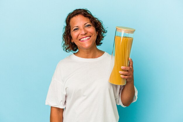 Lateinische Frau mittleren Alters, die Pasta-Glas auf blauem Hintergrund isoliert hält glücklich, lächelnd und fröhlich.