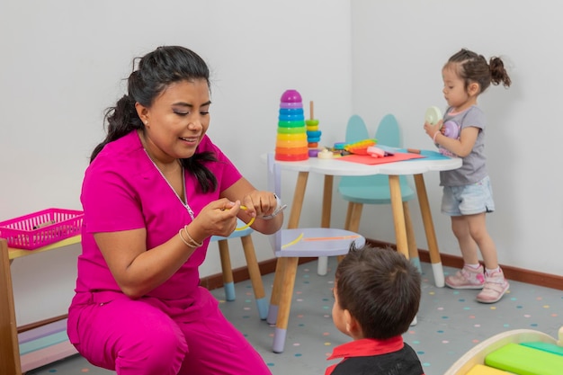 Lateinische Kinderärztin, die mit einem Kind in ihrer Arztpraxis spielt, während ein Mädchen auf einem Tisch im Hintergrund spielt