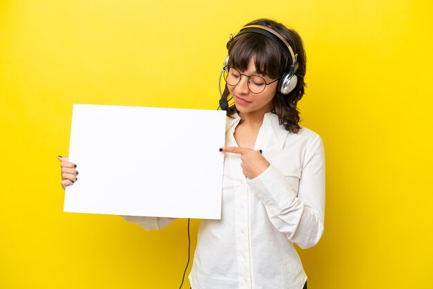 Lateinische Telemarketerin, die mit einem Headset arbeitet, isoliert auf gelbem Hintergrund, hält ein leeres Plakat mit fröhlichem Gesichtsausdruck und zeigt darauf