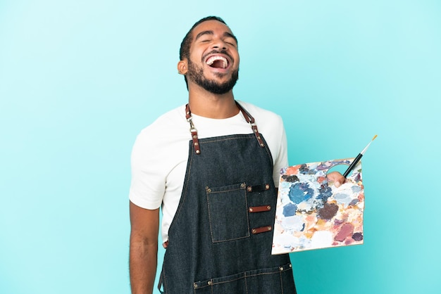 Lateinischer Mann des jungen Künstlers, der eine Palette lokalisiert auf blauem Hintergrund hält, lacht