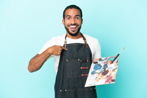 Foto lateinischer mann des jungen künstlers, der eine palette lokalisiert auf blauem hintergrund mit überraschtem gesichtsausdruck hält