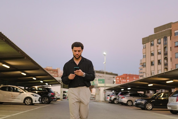 Lateinischer Mann geht in der Abenddämmerung mit einem Handy auf einem Parkplatz spazieren