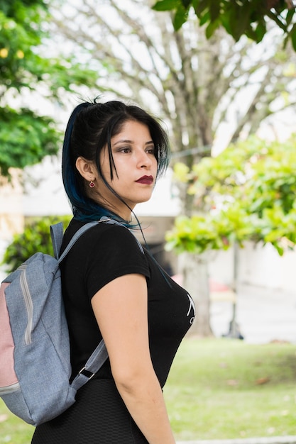 Lateinisches College-Mädchen mit blauen Haaren, das durch den Park geht, gekleidet in einer schwarzen und blauen Tasche, umgeben