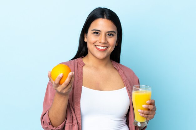 Latina jovem mulher isolada em azul, segurando uma laranja e um suco de laranja