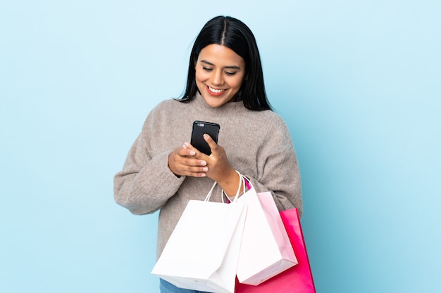 Latina jovem mulher isolada na parede azul segurando sacolas de compras e escrever uma mensagem com o celular para um amigo