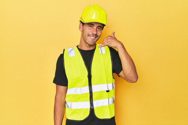 Foto latino-arbeiter in gelber weste und helm zeigt mit den fingern eine handyanruf-geste