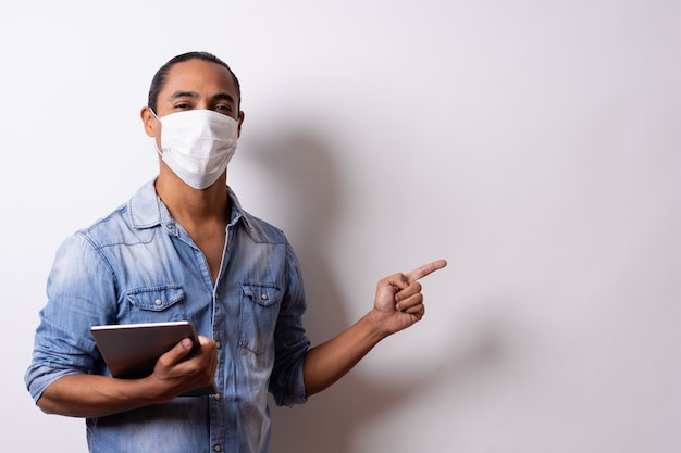 Latino Mann mit Gesichtsmaske hält eine Tablette und zeigt auf den leeren Raum auf weißem Hintergrund. Soziale Distanz.