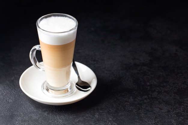 Latte Macchiato in einer Glastasse mit Henkel mit Kaffee herum und Löffel auf dunklem Hintergrund isoliert