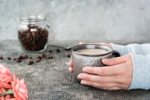 Latte en una taza de cerámica en manos femeninas un frasco de granos de café y rosas sobre la mesa