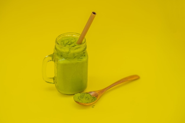 Latte de té verde con hielo en tarro de albañil y paja y cuchara con matcha en polvo sobre fondo amarillo Té de latte Matcha helado casero con leche cero desperdicio