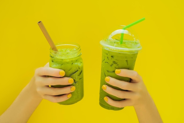 Latte de té verde con hielo en un tarro de albañil y en un vaso de plástico en las manos sobre fondo amarillo Té de latte matcha helado casero con leche cero desperdicio