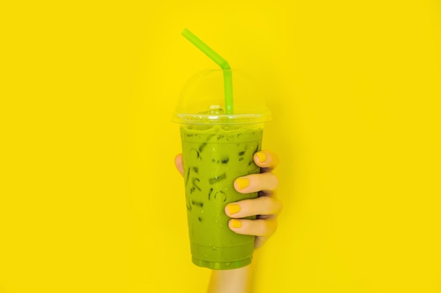 Latte de té verde con hielo en vaso de plástico y paja en mano femenina con manicura amarilla sobre fondo amarillo Té de latte matcha helado casero con leche para llevar