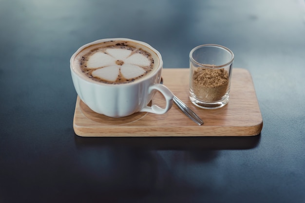 Lattekunstkaffee auf der weißen Schale heißem Kaffee auf hölzernem Behälter und dunkler Tabelle.