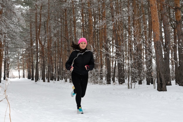 Laufsportfrau. Läuferin, die im kalten Winterwald joggt und warme sportliche Laufbekleidung und Kopfhörer trägt. Schöne Passform weibliches Fitness-Modell.